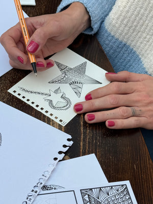 A close up of a hand using a clear Whimsy Lines acrylic template to draw organic flowing patterns in a sketchbook
